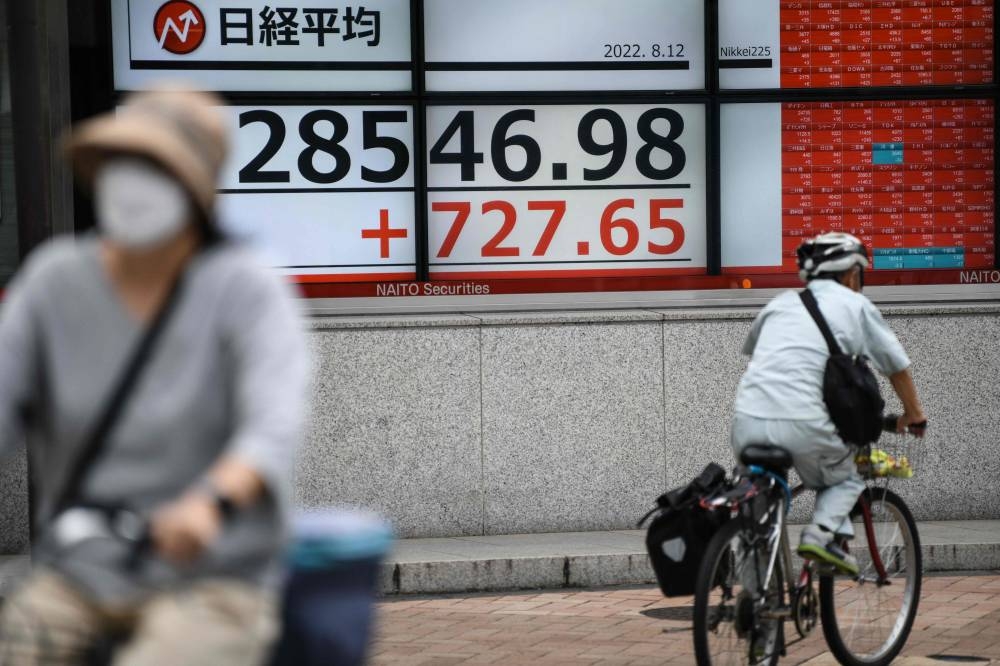 People cycle past a board showing the closing numbers on the Tokyo Stock Exchange in the business district of Tokyo on August 12, 2022. — AFP pic