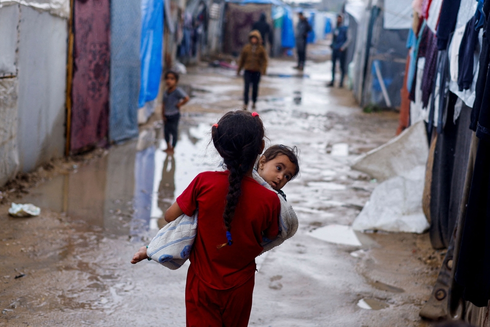A displaced Palestinian girl holds a child as she walks at a tent camp on a rainy day, amid the ongoing conflict between Israel and Hamas, in Rafah, in the southern Gaza Strip May 6, 2024. — Reuters pic