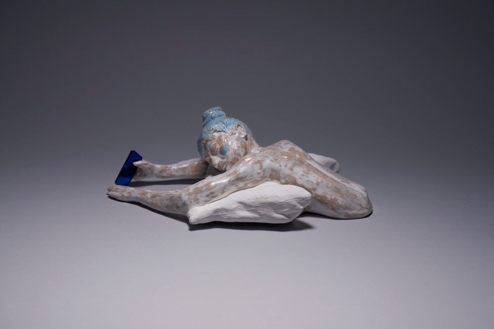 Tan’s ‘Letting Go’ ceramic sculpture depicts a ceramic figure laying on a pillow holding a mobile device emitting blue light.