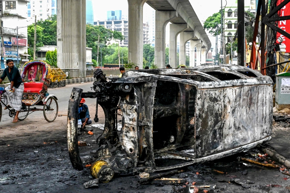 Passersby look at a burnt vehicle along a street amid anti-government protests in Dhaka on August 5, 2024. — AFP pic