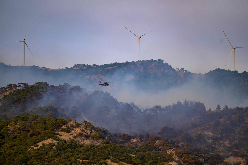 This photograph shows a helicopter carrying water to fight a forest fire, with windmills in the background in Turkey's western province of Izmir on August 17, 2024. — AFP pic