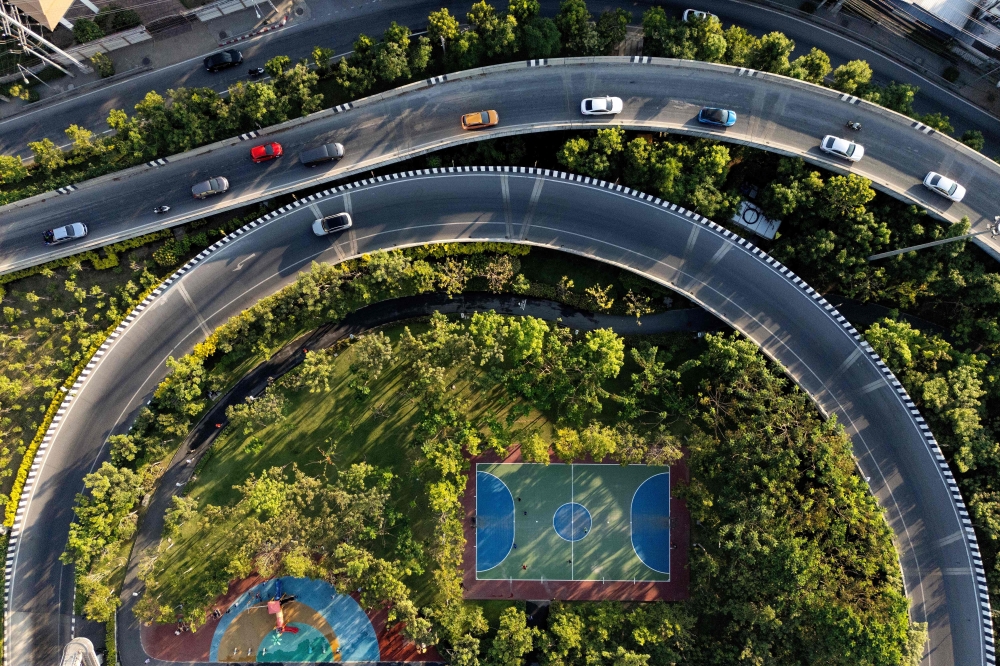 This aerial photograph taken on April 26, 2024 shows people playing football on a court at a pocket park next to a busy main road as evening rush-hour traffic drives past in Bangkok. A full tank here is more than double the cost of Malaysia’s petrol prices. — AFP pic