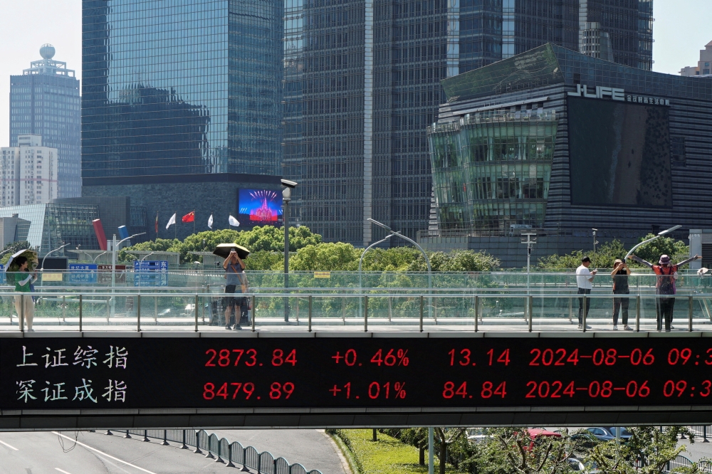 People take pictures on an overpass with a display of stock information in front of buildings in the Lujiazui financial district in Shanghai, China August 6, 2024. — Reuters pic